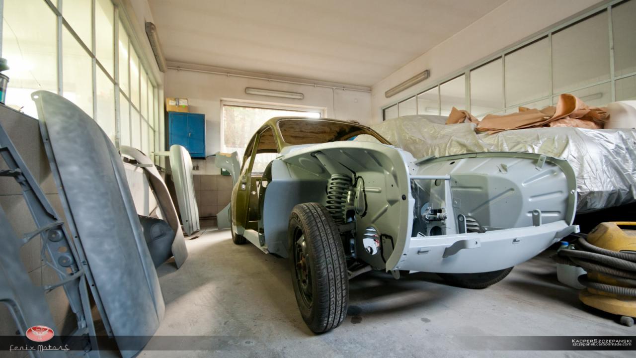 Klasyczny samochód w trakcie renowacji w warsztacie, z widocznymi elementami karoserii przygotowanymi do lakierowania, odsłoniętym zawieszeniem i częściowo zdemontowanym wnętrzem. Dodatkowe części...