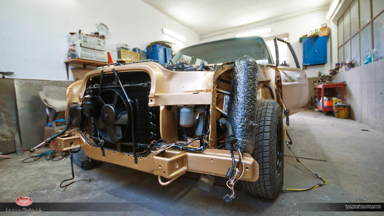 Klasyczny samochód w trakcie renowacji w warsztacie, z widoczną zdemontowaną karoserią, odsłoniętym silnikiem i elementami podwozia, częściowo pomalowany na złoty kolor, z widocznymi narzędziami...