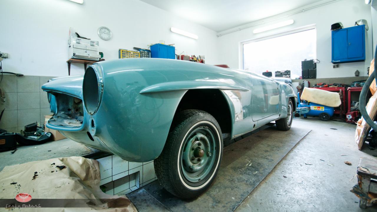 Turkusowy, klasyczny roadster bez przedniego zderzaka i reflektorów, na podnośniku w warsztacie, widoczne narzędzia i wyposażenie warsztatowe w tle.