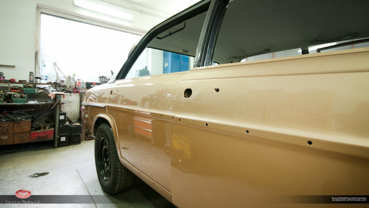 Bok beżowego samochodu po usunięciu elementów karoserii, widoczne otwory montażowe, w tle warsztat z narzędziami i oknem.