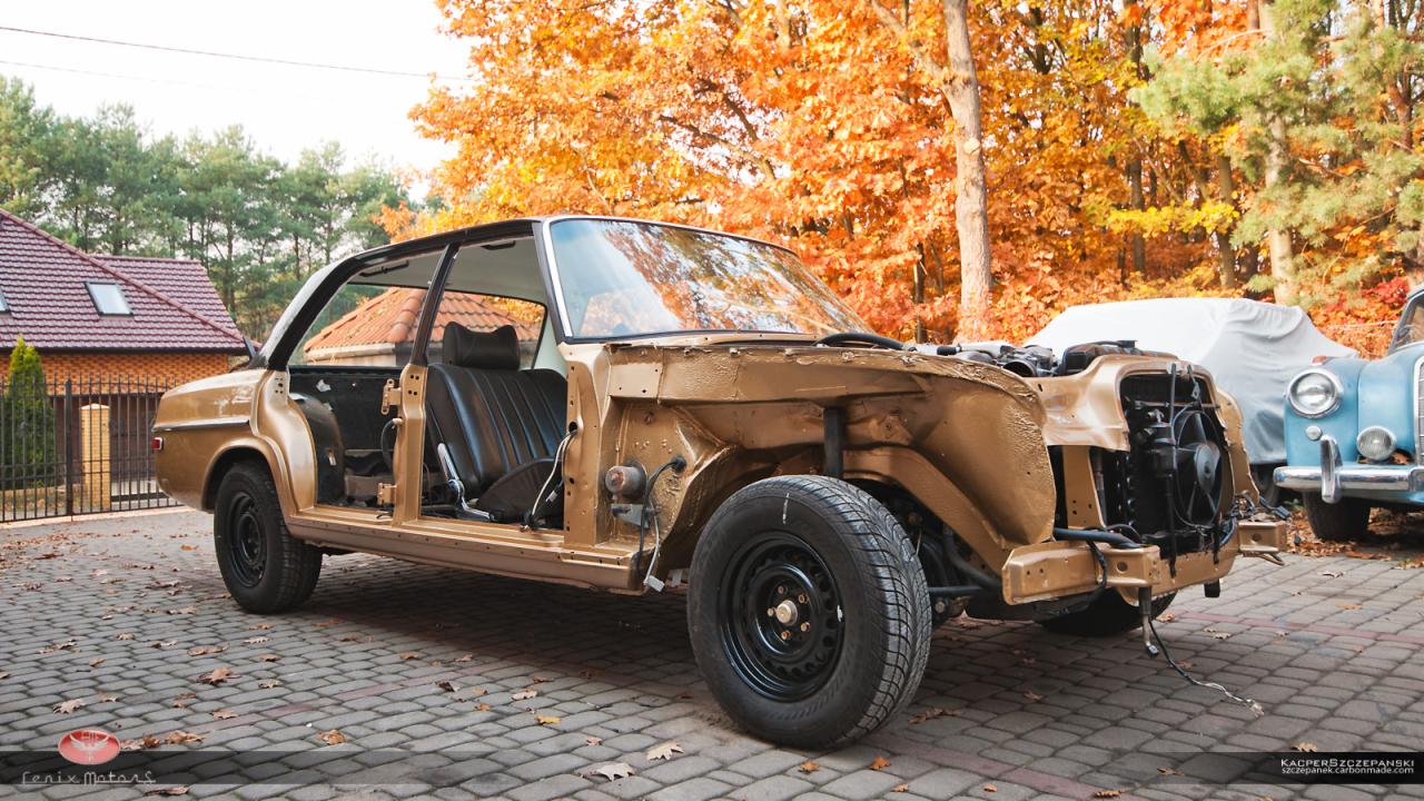 Klasyczny samochód Mercedes w trakcie renowacji blacharskiej, widoczne elementy karoserii w kolorze złotym, brak drzwi i maski, na pierwszym planie czarne koło, w tle jesienne drzewa i budynek...