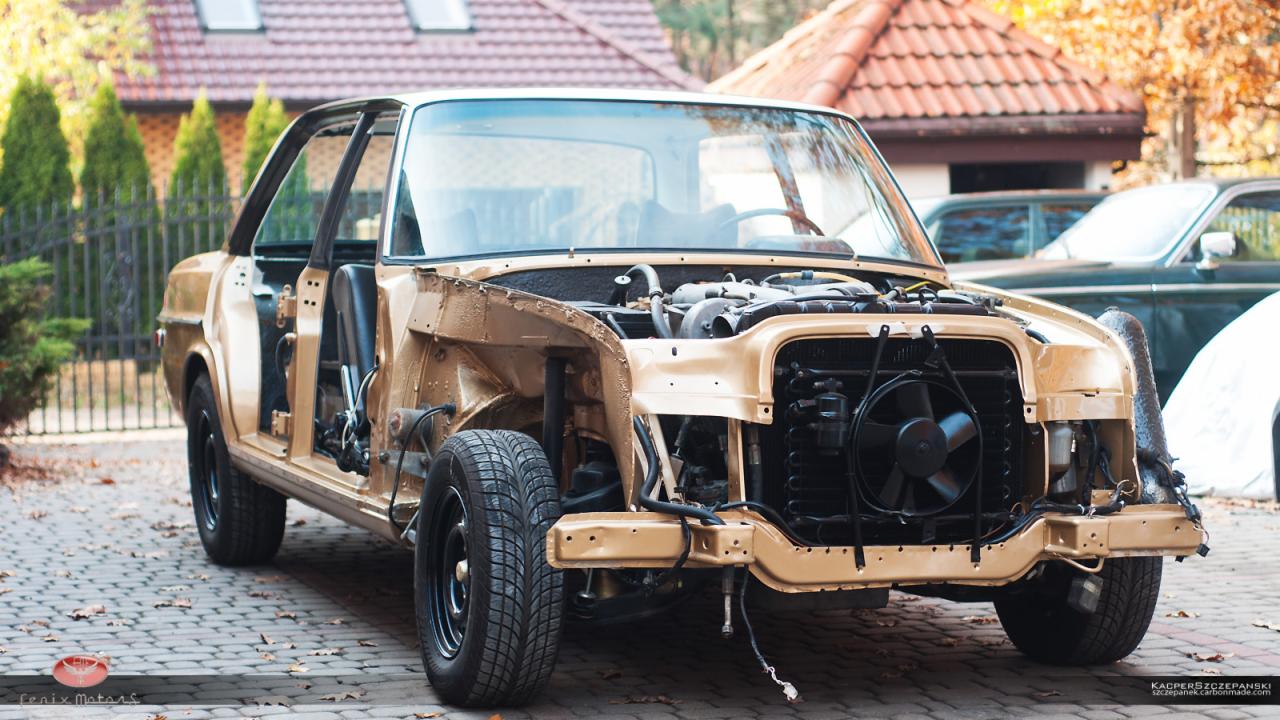 Klasyczny samochód w trakcie renowacji, z częściowo zdemontowanym nadwoziem i widocznym silnikiem, zaparkowany na brukowanym podjeździe w jesiennej scenerii.