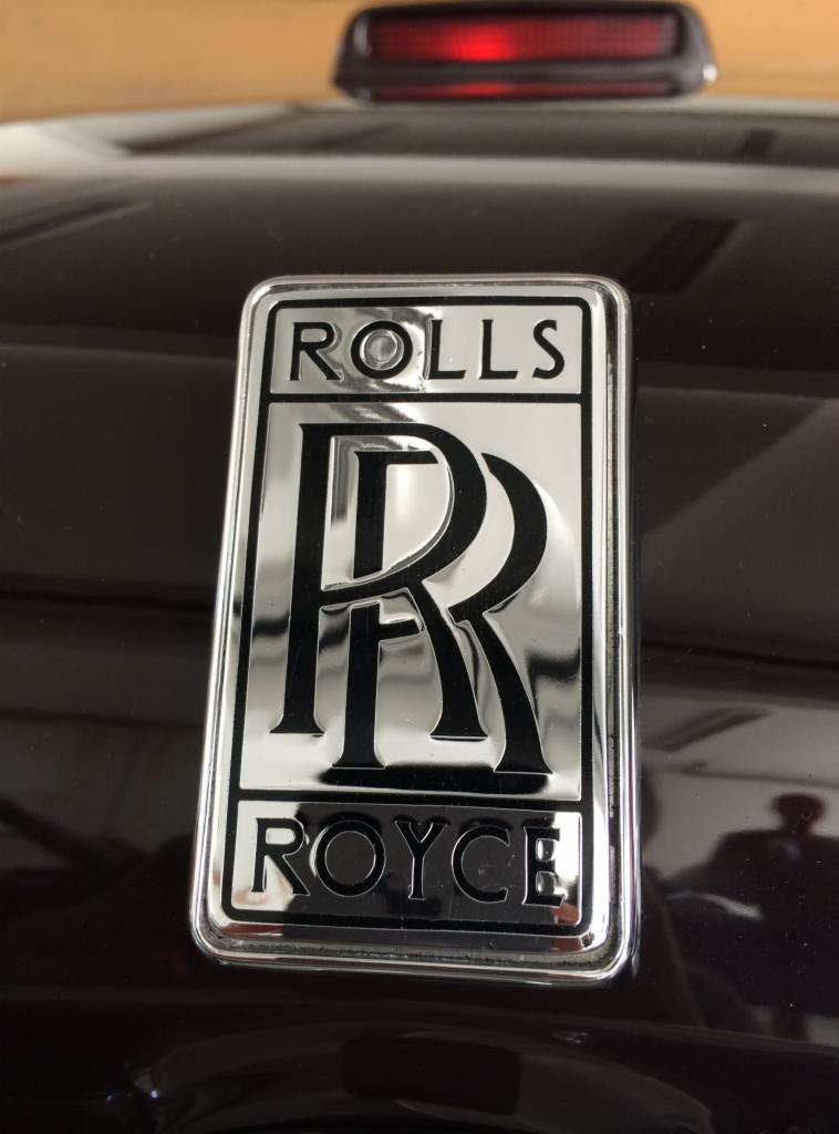 Błyszczący, chromowany emblemat Rolls-Royce na ciemnym lakierze samochodu, widoczny fragment trzeciego światła stop oraz odbicie sylwetki fotografa.