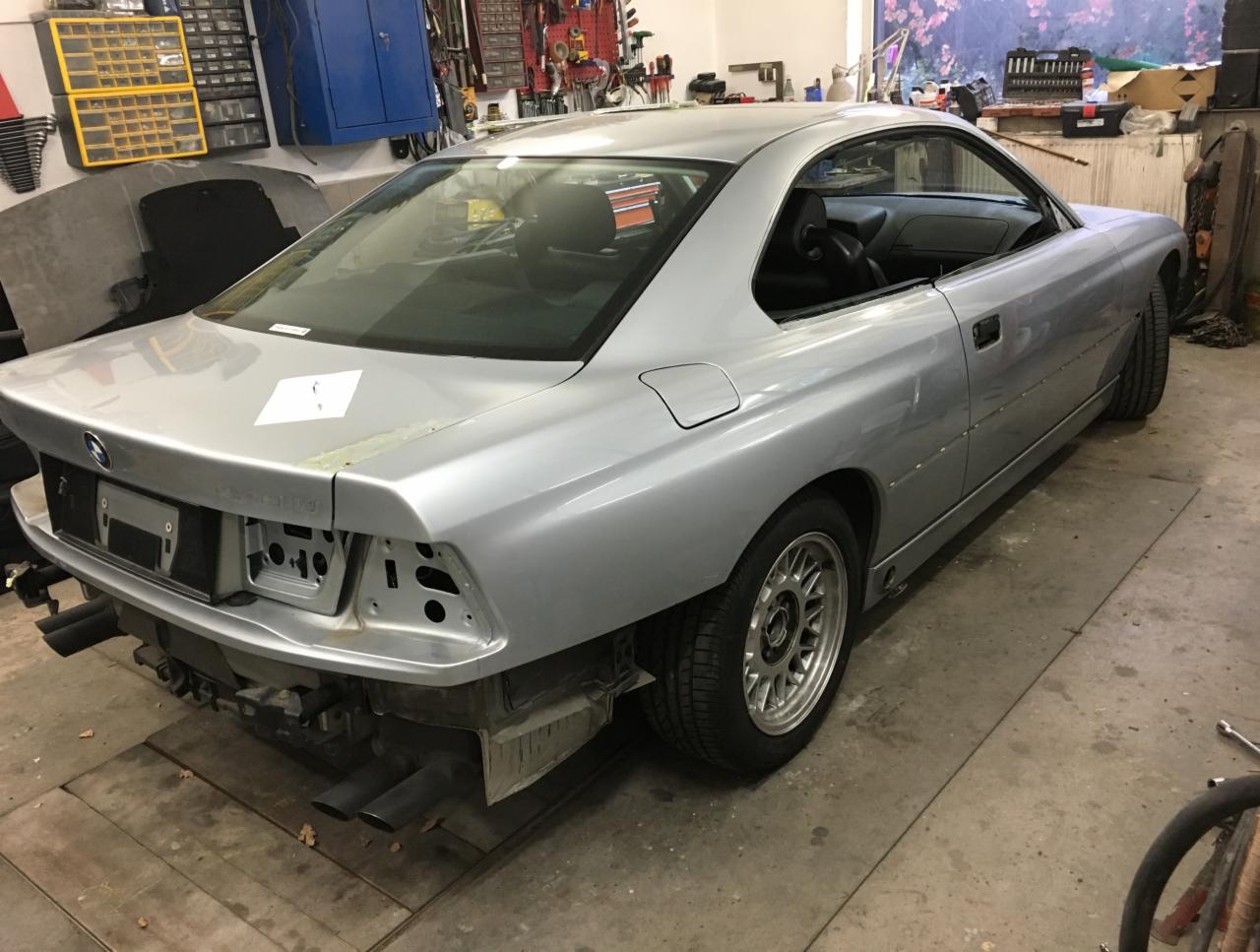 Srebrne BMW 850i bez tylnego zderzaka w warsztacie, widoczne elementy blacharskie w trakcie naprawy, naklejka na klapie bagażnika, narzędzia i szafki w tle.