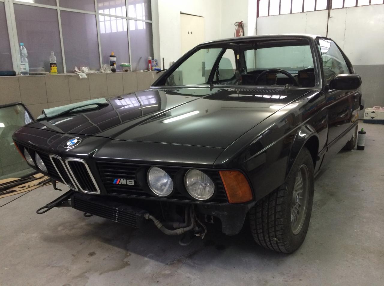 Czarny, klasyczny model BMW M6 bez przedniego zderzaka, w trakcie prac blacharskich w warsztacie, widoczna ściereczka na masce, reflektory i atrapa chłodnicy, wnętrze warsztatu z oknami...