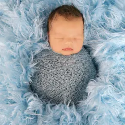 Śpiący noworodek zawinięty w niebieski kocyk na tle błękitnego, puszystego materiału.