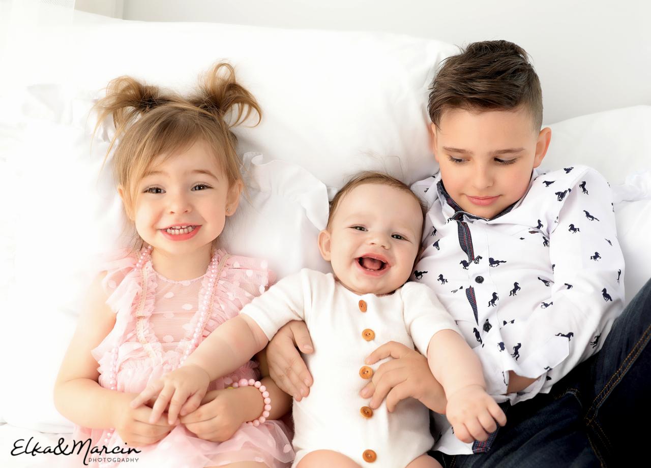 Uśmiechnięta dziewczynka w różowej sukience, niemowlę w białym body z brązowymi guzikami i chłopiec w koszuli w konie leżą na białym tle.