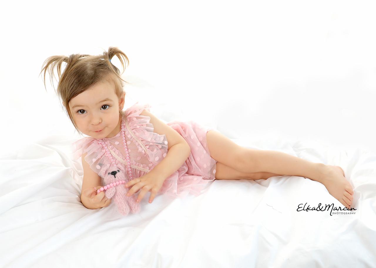 Dziewczynka w różowej sukience z falbankami i warkoczykami leży na białym tle, trzymając pluszowego misia z koralikami.