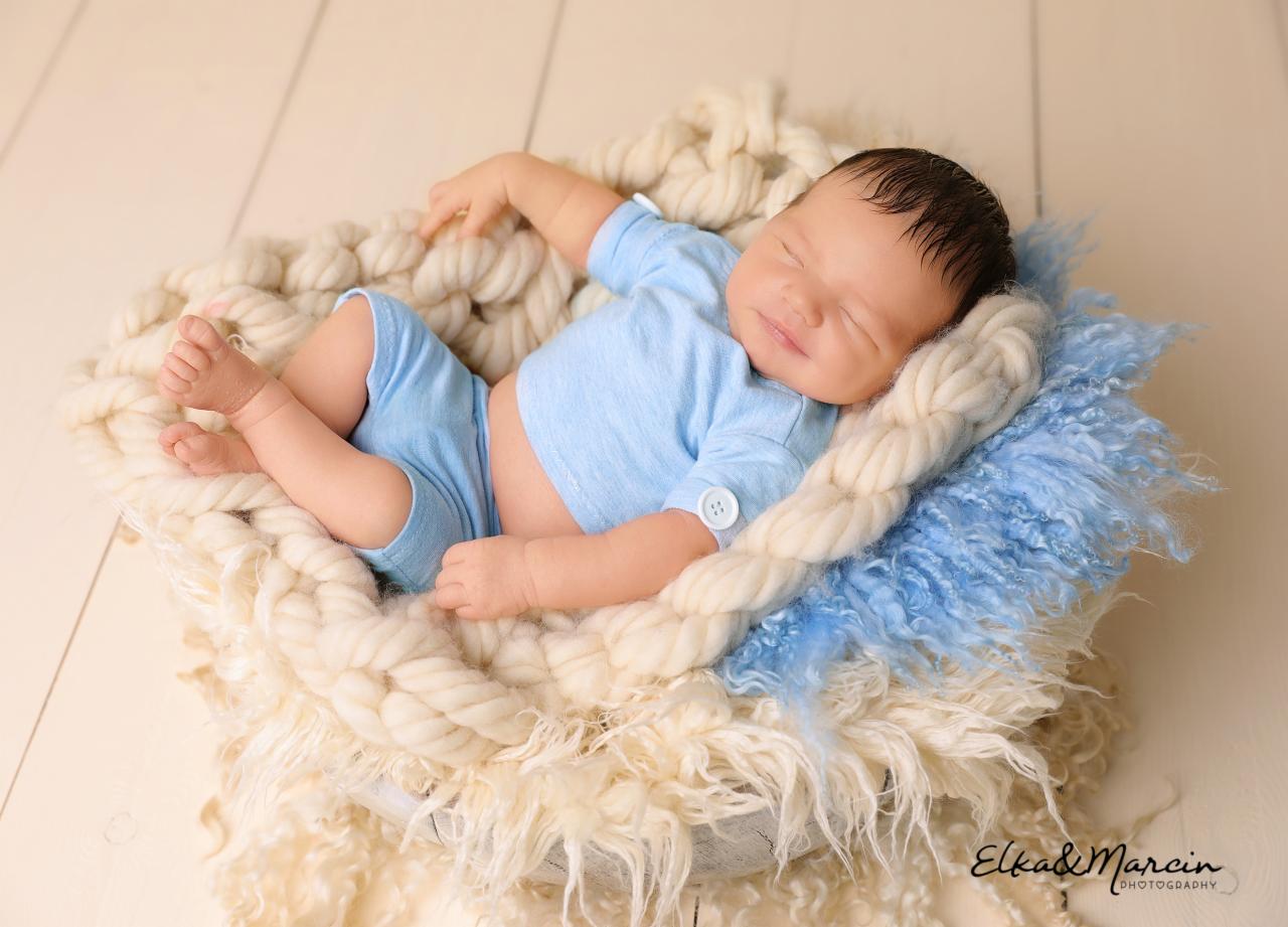 Śpiący noworodek w błękitnym ubranku, ułożony w koszyku wyłożonym kremową plecionką i niebieskim futerkiem, na tle drewnianej podłogi.