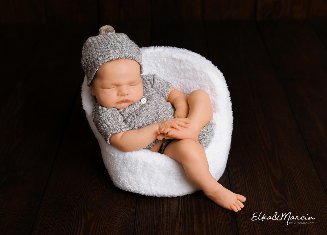 Śpiący noworodek w szarym, dzierganym komplecie i czapeczce z pomponem, ułożony w białym, pluszowym foteliku na ciemnym, drewnianym tle.