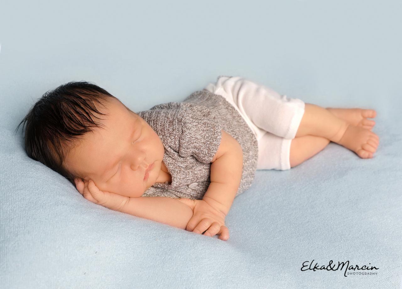 Śpiący noworodek ubrany w szary sweterek i białe spodenki, leżący na błękitnym tle, z delikatnie podpartą głową na dłoni.