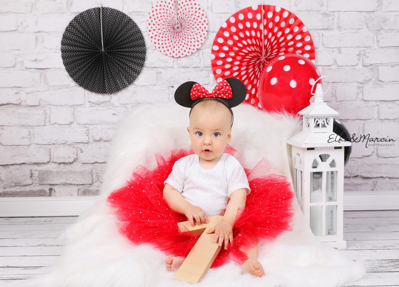 Urocze niemowlę w czerwonej tiulowej spódniczce i opasce z uszami Myszki Minnie, siedzące na białym futrze z drewnianą cyfrą '1', na tle ceglanej ściany z dekoracjami w kropki i białą latarnią.