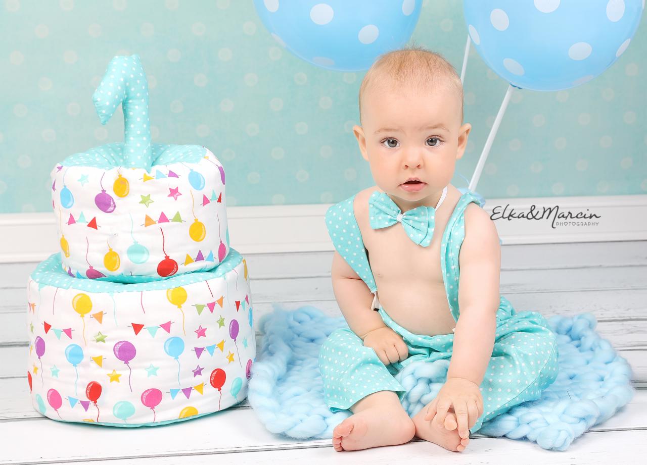 Uroczy niemowlak w błękitnym stroju z kokardą siedzi na puszystym kocyku obok dwupoziomowego tortu z materiału ozdobionego balonami i cyfrą '1', błękitne balony w tle.