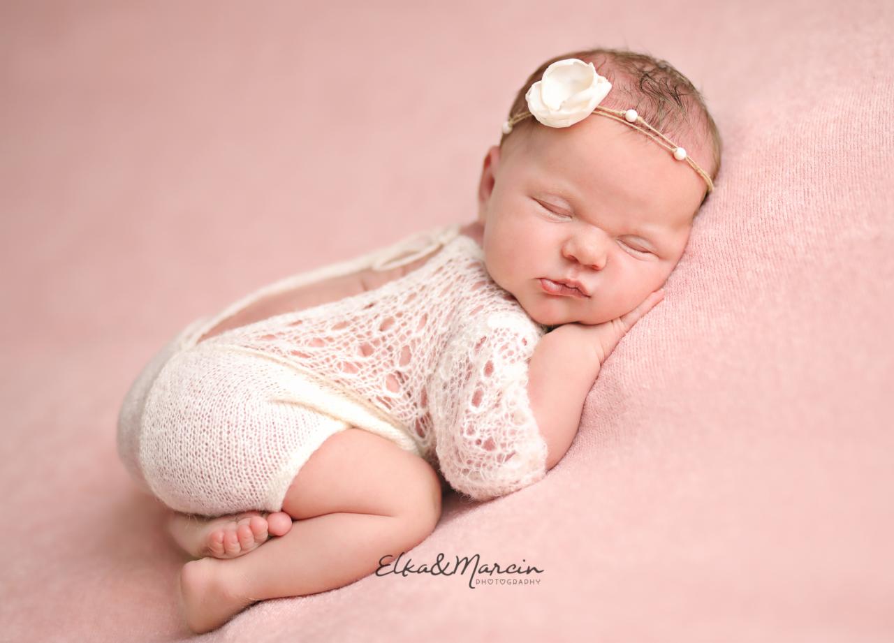 Śpiące niemowlę w ażurowym ubranku z opaską z białym kwiatkiem na różowym tle.