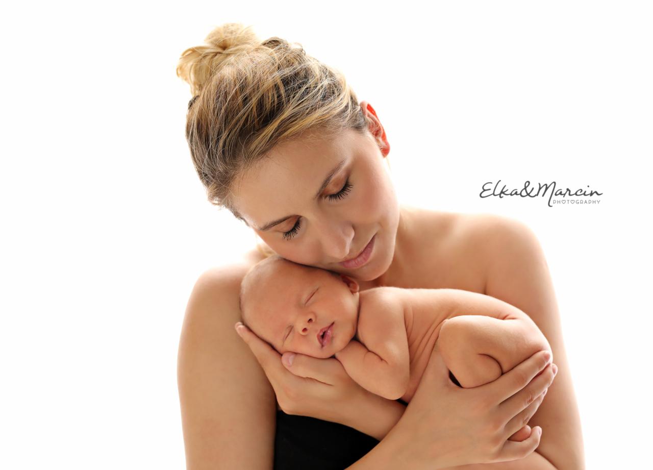 Matka z blond włosami upiętymi w kok, trzymająca na rękach śpiącego noworodka na jasnym tle, widoczne logo fotografa Elka&Marcin.