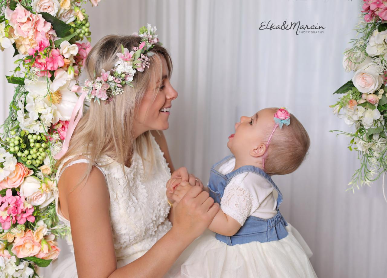 Uśmiechnięta blondynka w kwiecistym wianku trzyma za ręce małą dziewczynkę z różową opaską na głowie, na tle białej zasłony i kompozycji z różowych i białych kwiatów.