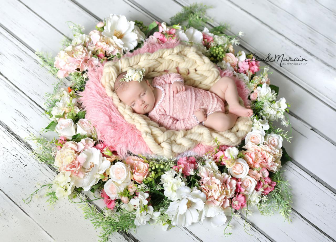 Śpiąca noworodka w różowym ubranku leży na puszystym kocyku w gnieździe z grubego warkocza, otoczonym kompozycją z różowych i białych kwiatów na tle drewnianych desek.