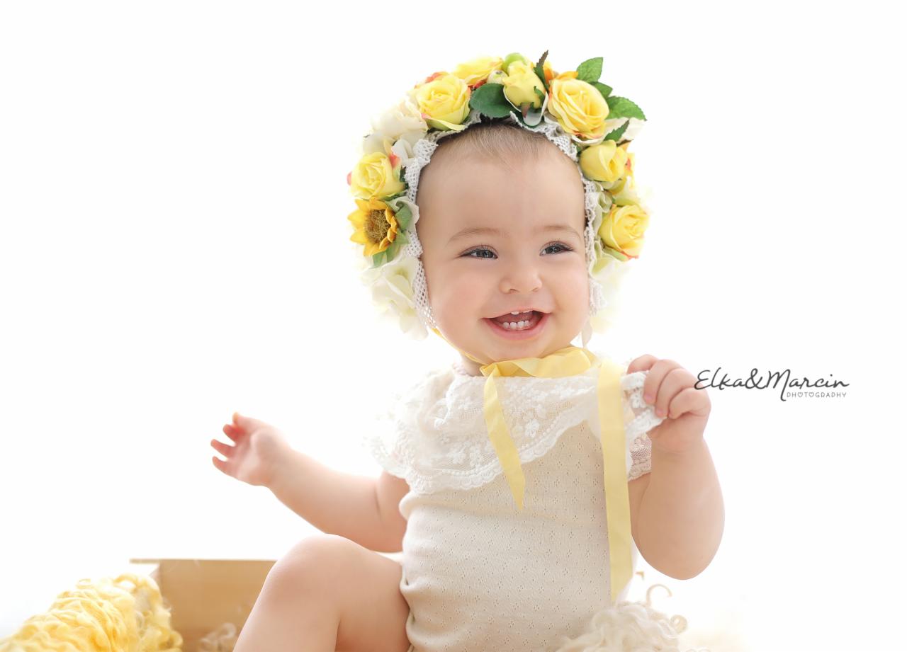Uśmiechnięta niemowlęca dziewczynka w koronie z żółtych róż i słonecznika, ubrana w kremowe body z koronkowym kołnierzykiem, siedząca w drewnianym pudełku z żółtą wełną.