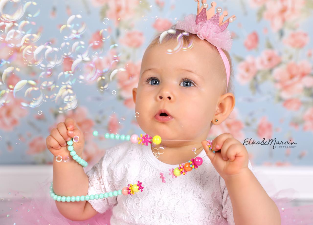 Niemowlę z niebieskimi oczami, w koronie i białej bluzce, otoczone bańkami mydlanymi, trzymające kolorowy naszyjnik na tle kwiatowego wzoru.