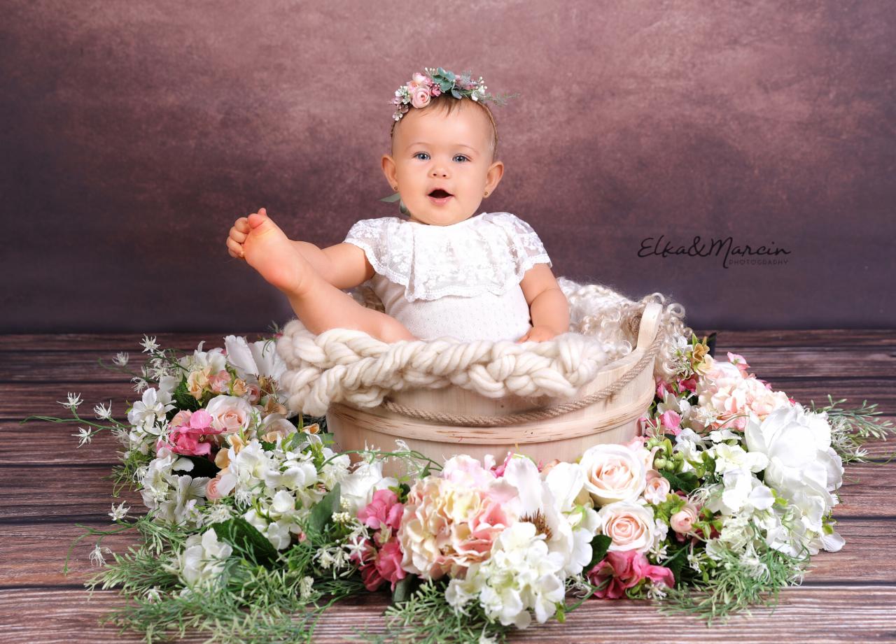 Niemowlę w białym ubranku i wianku na głowie siedzi w drewnianym wiaderku wyłożonym grubym, kremowym sznurem, otoczonym kompozycją z różowych i białych kwiatów na drewnianym tle.