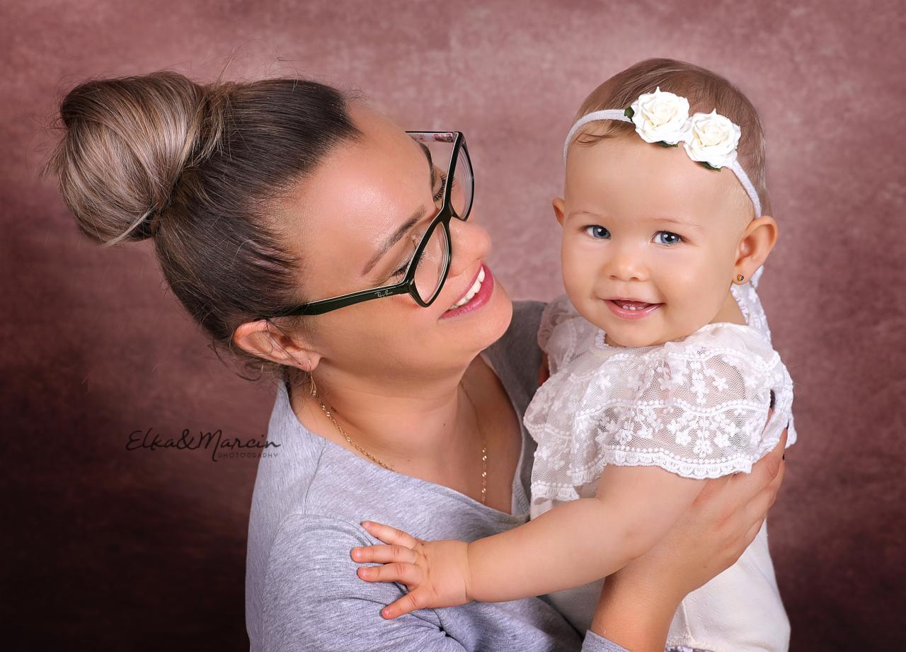 Uśmiechnięta matka w okularach trzyma na rękach ubraną w białą koronkę dziewczynkę z opaską z białych kwiatów na głowie, na rozmytym różowym tle.