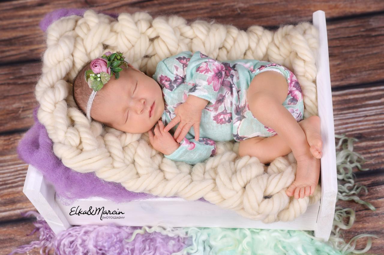 Śpiąca noworodka w pastelowym kombinezonie z kwiatowym wzorem, na głowie opaska z różowymi kwiatami, ułożona w małym białym łóżeczku wyłożonym grubym, kremowym pledem z warkoczami, na tle...