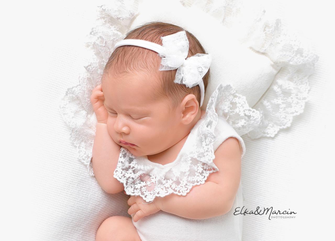 Śpiąca noworodka w białym ubranku z koronkowym wykończeniem i opaską z kokardką na głowie, leżąca na białym, teksturowanym kocyku z koronkową obwódką, delikatne oświetlenie.