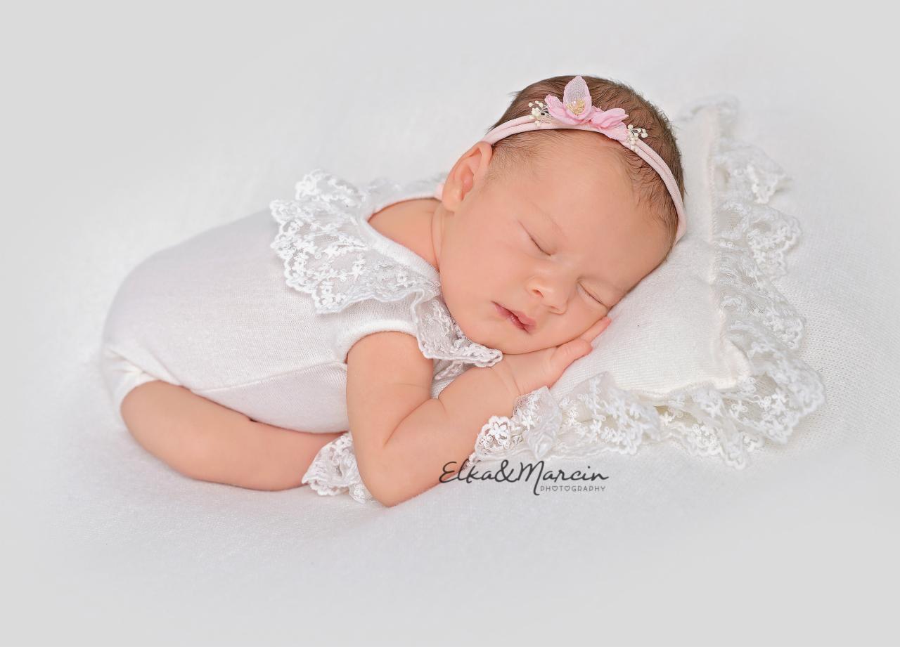 Śpiąca noworodka w białym body z koronkowym kołnierzykiem i opaską z różowym kwiatkiem na białej poduszce z koronkową falbanką, zdjęcie z bliska.