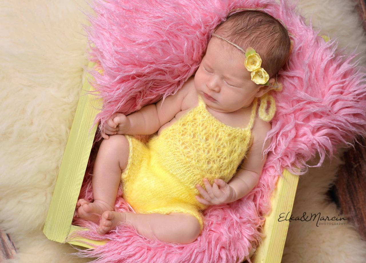 Śpiąca noworodka w żółtym stroju, z kwiatową opaską, ułożona w pastelowo-różowym futerku wewnątrz jasnożółtej drewnianej ramki.