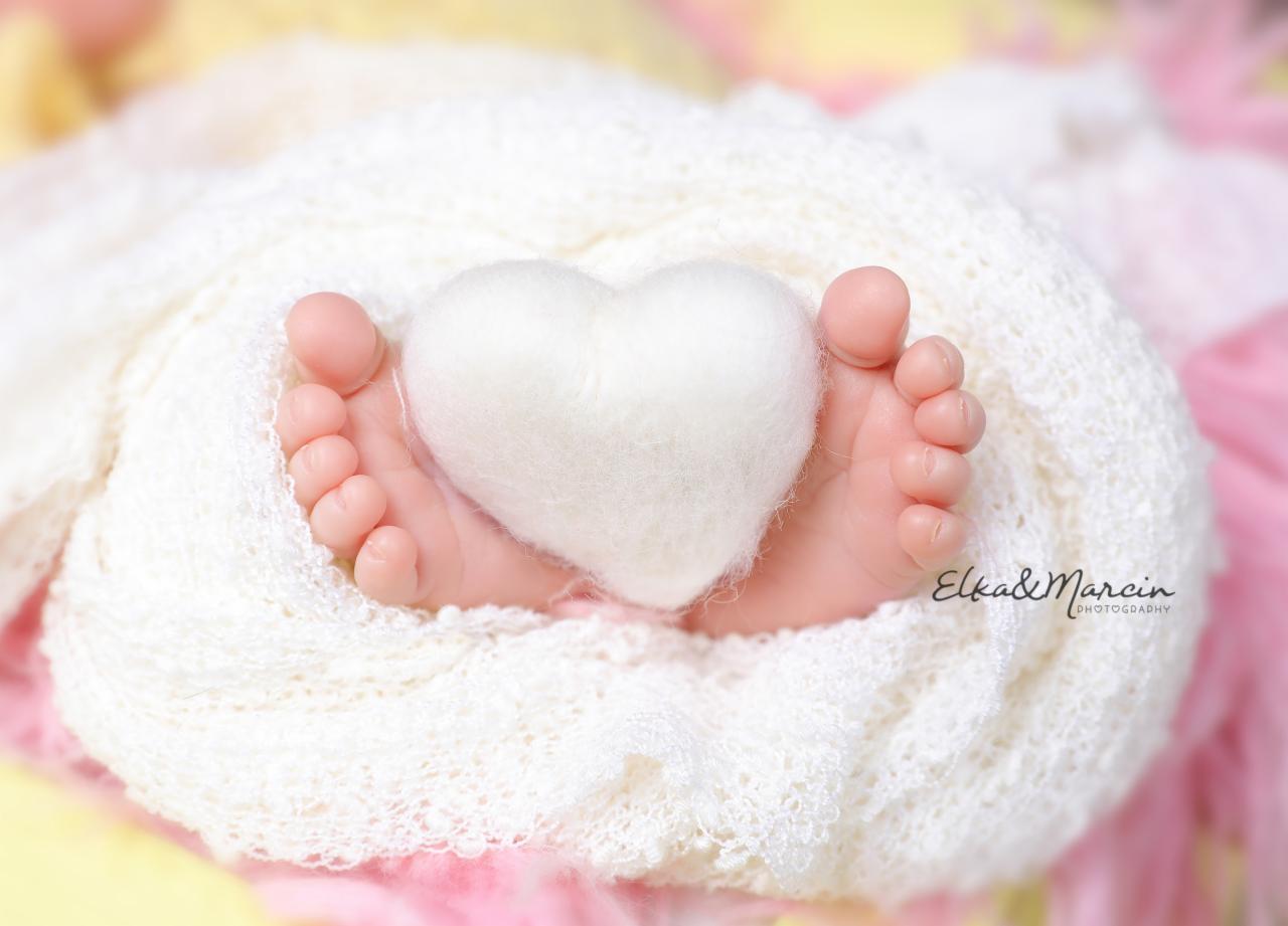 Malutkie stopy noworodka otulone kremowym kocykiem, z białym, filcowym sercem pośrodku, delikatne pastelowe tło, logo fotografa w prawym dolnym rogu.