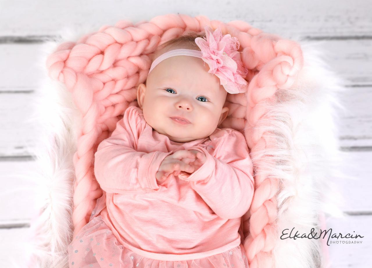 Uśmiechnięta niemowlęca dziewczynka w różowej sukience i opasce z kwiatem, leżąca w koszyku wyłożonym puszystym futerkiem i grubym różowym kocem na tle białych desek.