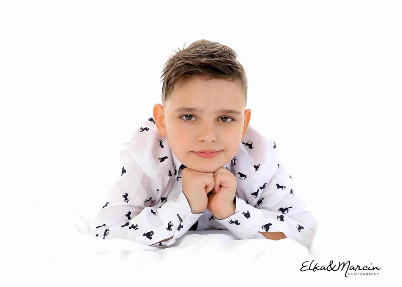 Portret chłopca leżącego na białym tle, w koszuli z motywem koni, dłonie pod brodą, delikatny uśmiech, z logo fotografa w prawym dolnym rogu.