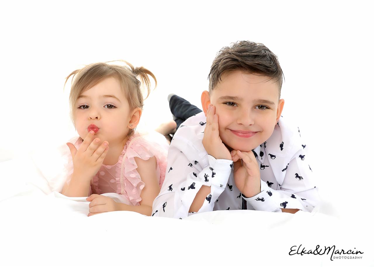 Portret rodzeństwa leżącego na białym tle: dziewczynka w różowej sukience robi buziaka, a chłopiec w koszuli w konie uśmiecha się, opierając policzek na dłoni.