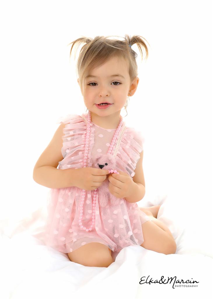 Dziewczynka w różowej sukience i z różowym naszyjnikiem z pereł trzyma pluszowego misia na białym tle.