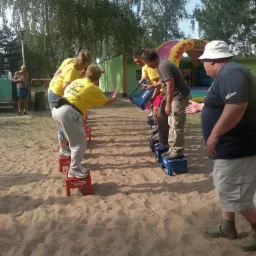 Grupa osób uczestniczy w konkurencji chodzenia po skrzynkach na piasku, z dmuchaną zjeżdżalnią w tle, podczas imprezy plenerowej.