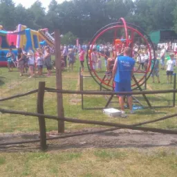 Plenerowa impreza z dmuchańcami, trampoliną i karuzelą dla dzieci, ogrodzona niskim płotem z gałęzi, w tle tłum ludzi i las.