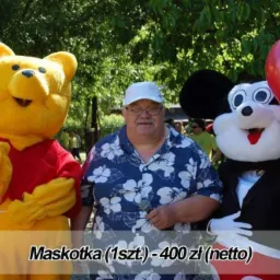 Mężczyzna w koszuli w kwiaty stoi pomiędzy maskotkami Kubusia Puchatka z żółtym balonem i Myszki Miki w garniturze trzymającej balony, na tle zieleni.