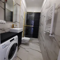 Wąska łazienka w bieli i czerni z marmurowymi płytkami, złotymi akcentami, białą pralką Candy, umywalką nablatową i grzejnikiem drabinkowym.
