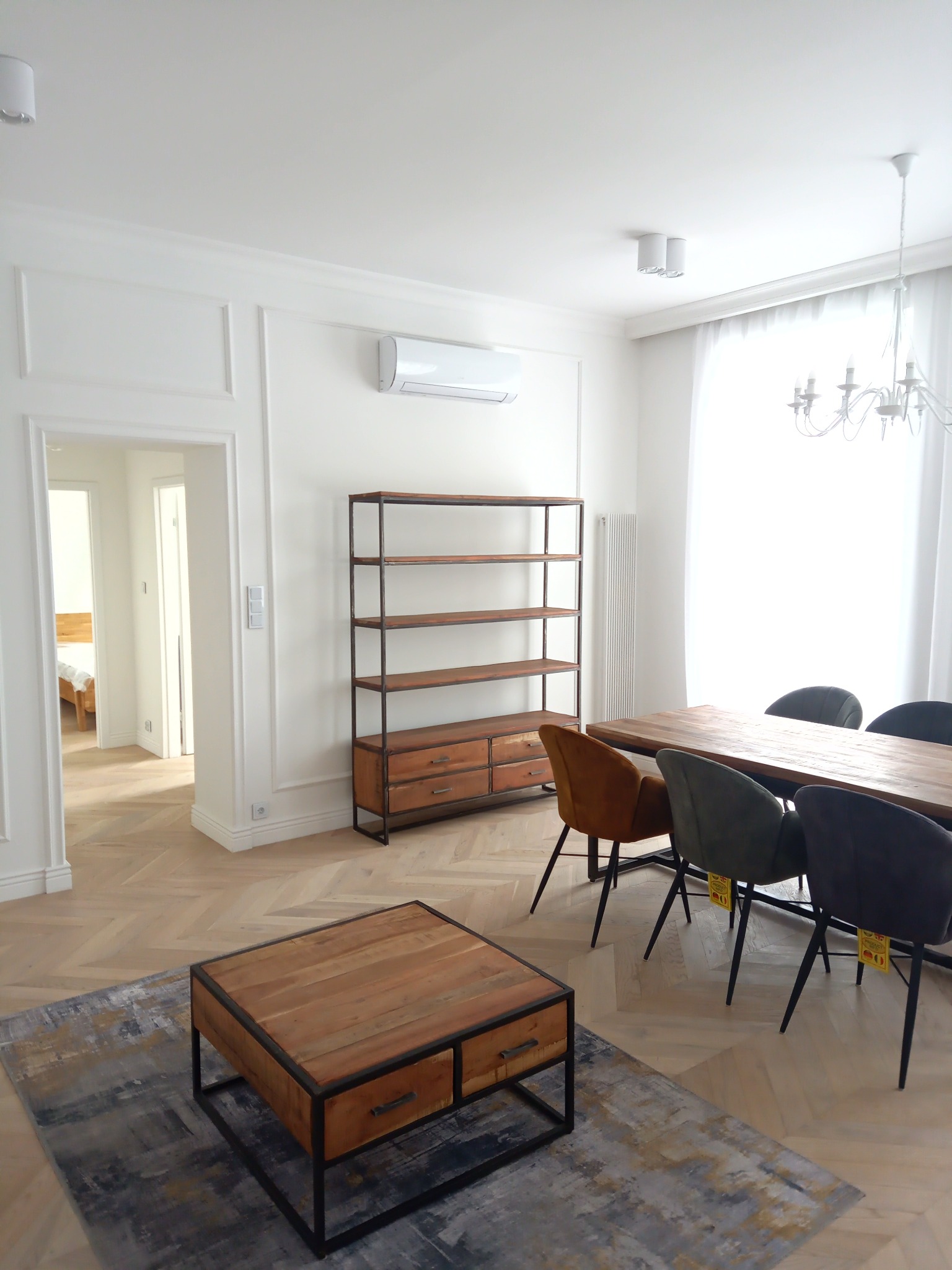 Jasne, przestronne wnętrze z drewnianą podłogą ułożoną w jodełkę, meblami w stylu industrialnym (regał, stolik kawowy), długim drewnianym stołem i krzesłami w odcieniach szarości i brązu, widok...