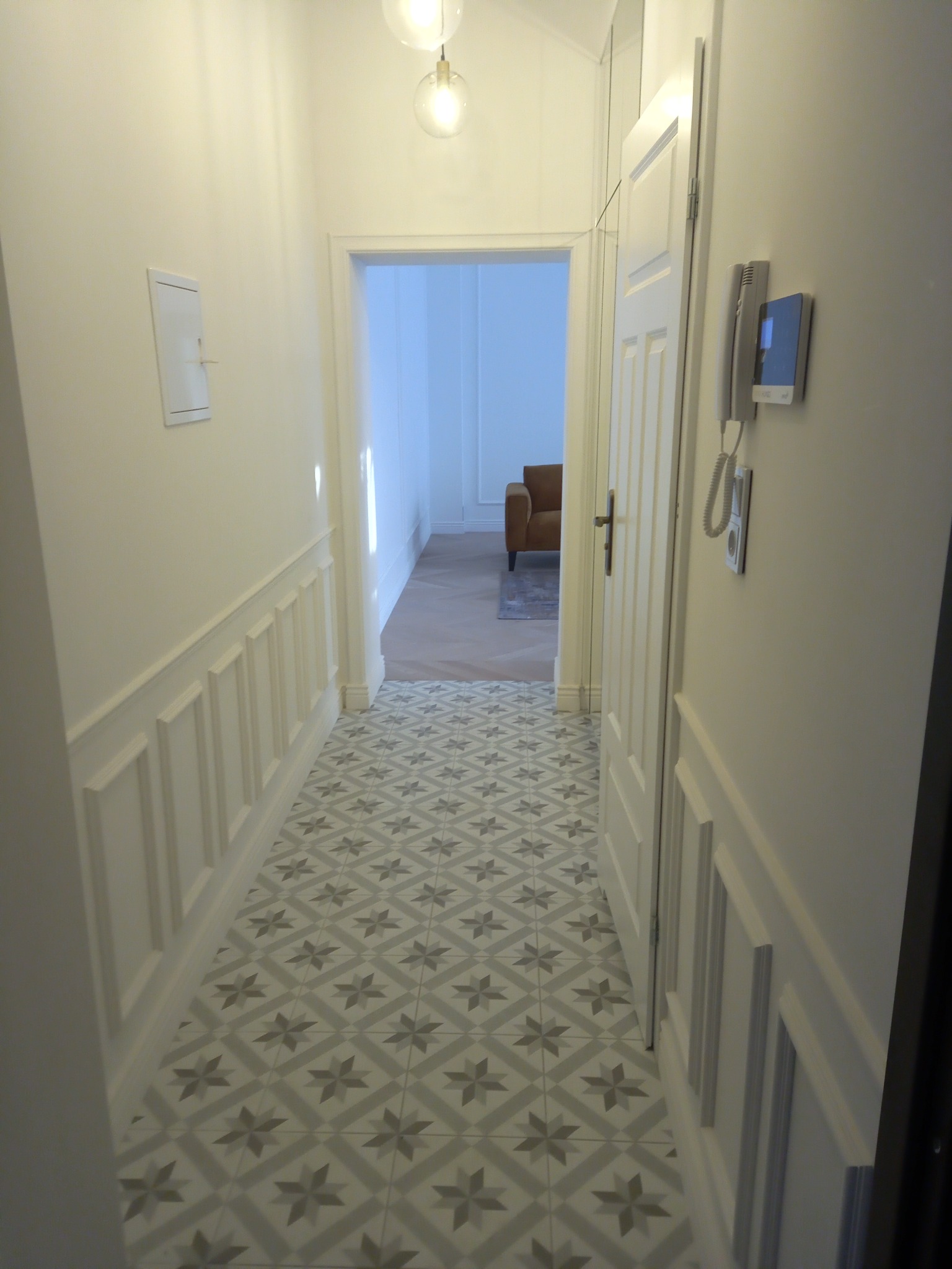 Korytarz po remoncie z panelami ściennymi, wzorzystą podłogą i widocznym fragmentem salonu z brązowym fotelem w głębi.