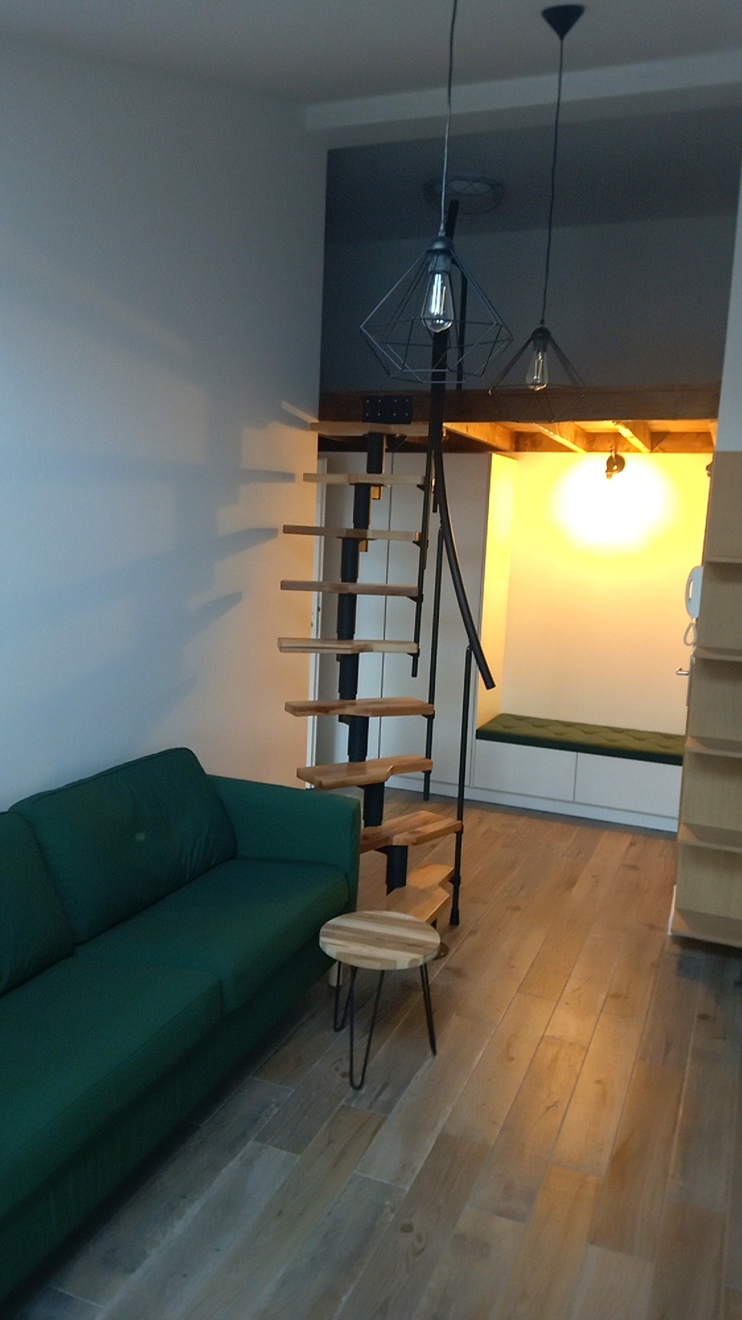 Spiralne schody z drewnianymi stopniami i czarną metalową konstrukcją w nowocześnie urządzonym mieszkaniu z zieloną sofą i małym stolikiem.