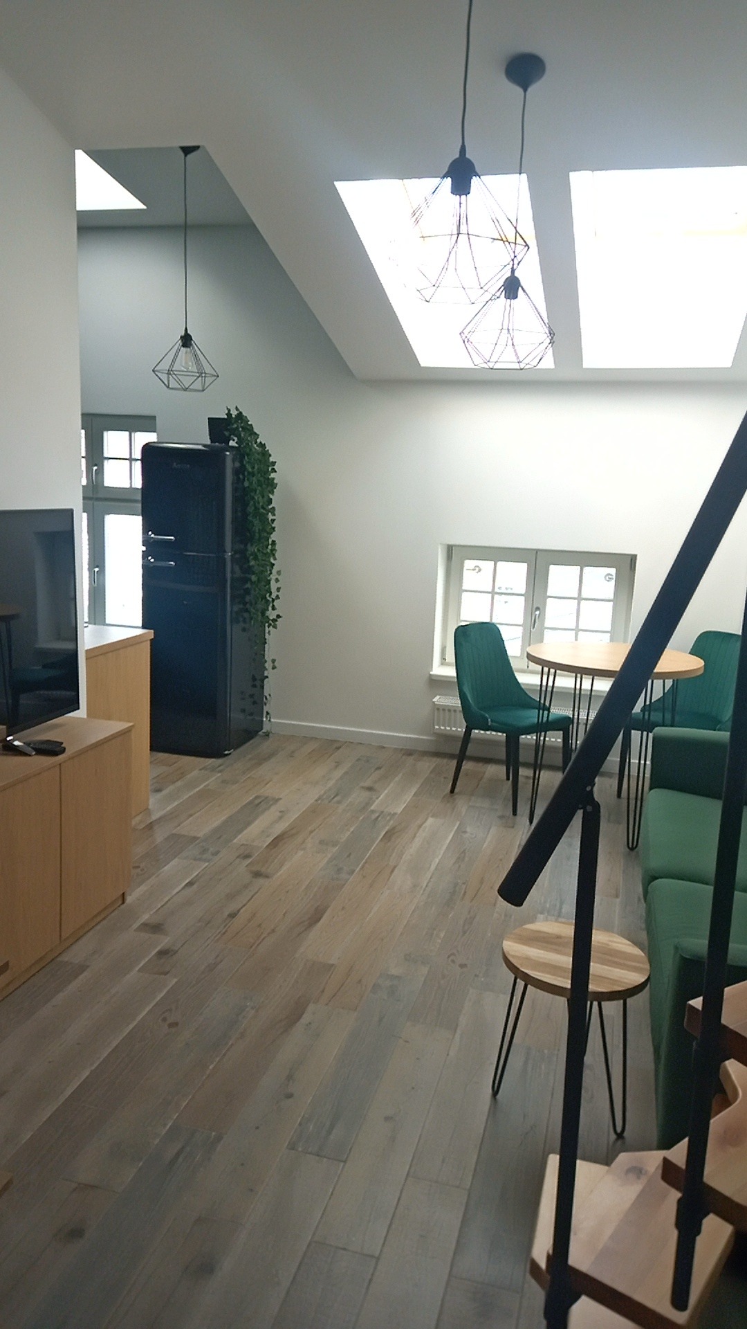 Jasne wnętrze z drewnianą podłogą, czarną lodówką w stylu retro, zielonymi krzesłami i sofą, oświetlone przez okna dachowe i lampy wiszące o geometrycznych kształtach.