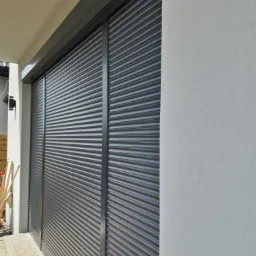 Zewnętrzna, antracytowa osłona okienna z lamelami, zamontowana na elewacji budynku z jasnoszarym tynkiem strukturalnym.