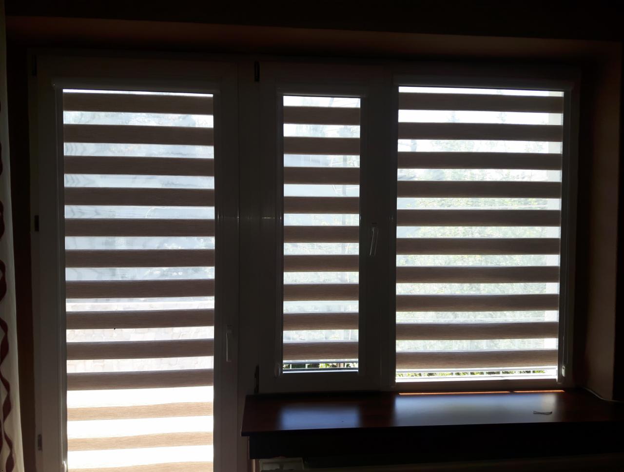 Trzy okna z zamontowanymi poziomymi przesłonami okiennymi, częściowo zasłoniętymi, w pomieszczeniu o ciemnych ścianach.