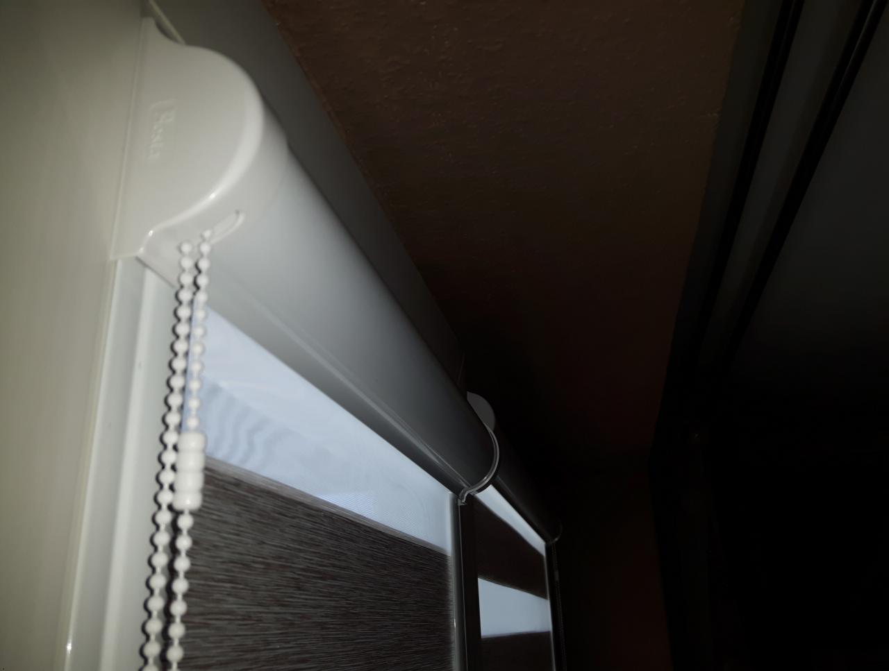 Fragment okna z zamontowaną, dwuwarstwową przesłoną okienną z białym mechanizmem łańcuszkowym, widok z dołu.