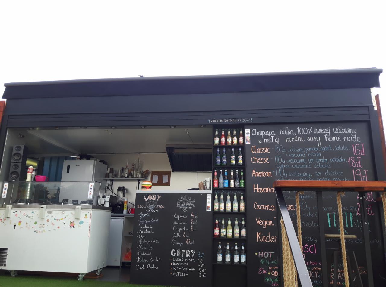Otwarta, czarna przyczepa gastronomiczna z menu wypisanym kredą na tablicach: lody, kawa, gofry, burgery, napoje; w tle widoczne wyposażenie kuchni.