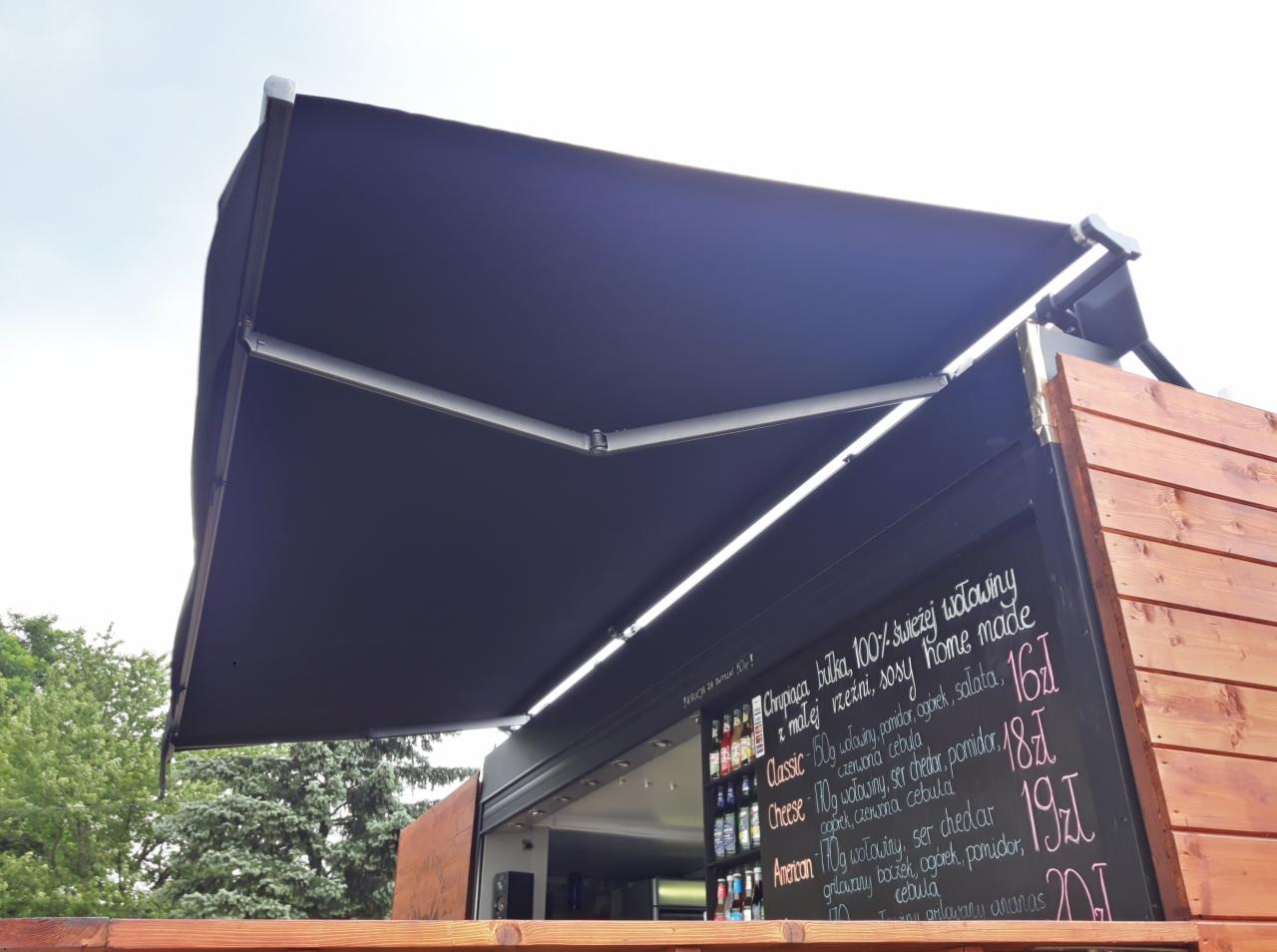 Zewnętrzna, rozkładana, czarna osłona przeciwsłoneczna nad food truckiem z menu wypisanym kredą.