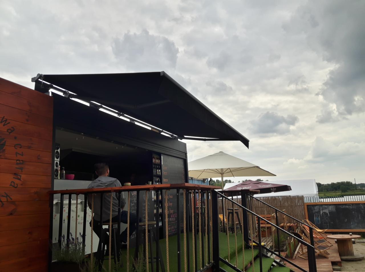 Czarny, rozkładany daszek nad kontenerem gastronomicznym z widocznym menu na tablicy i klientem siedzącym przy barze.