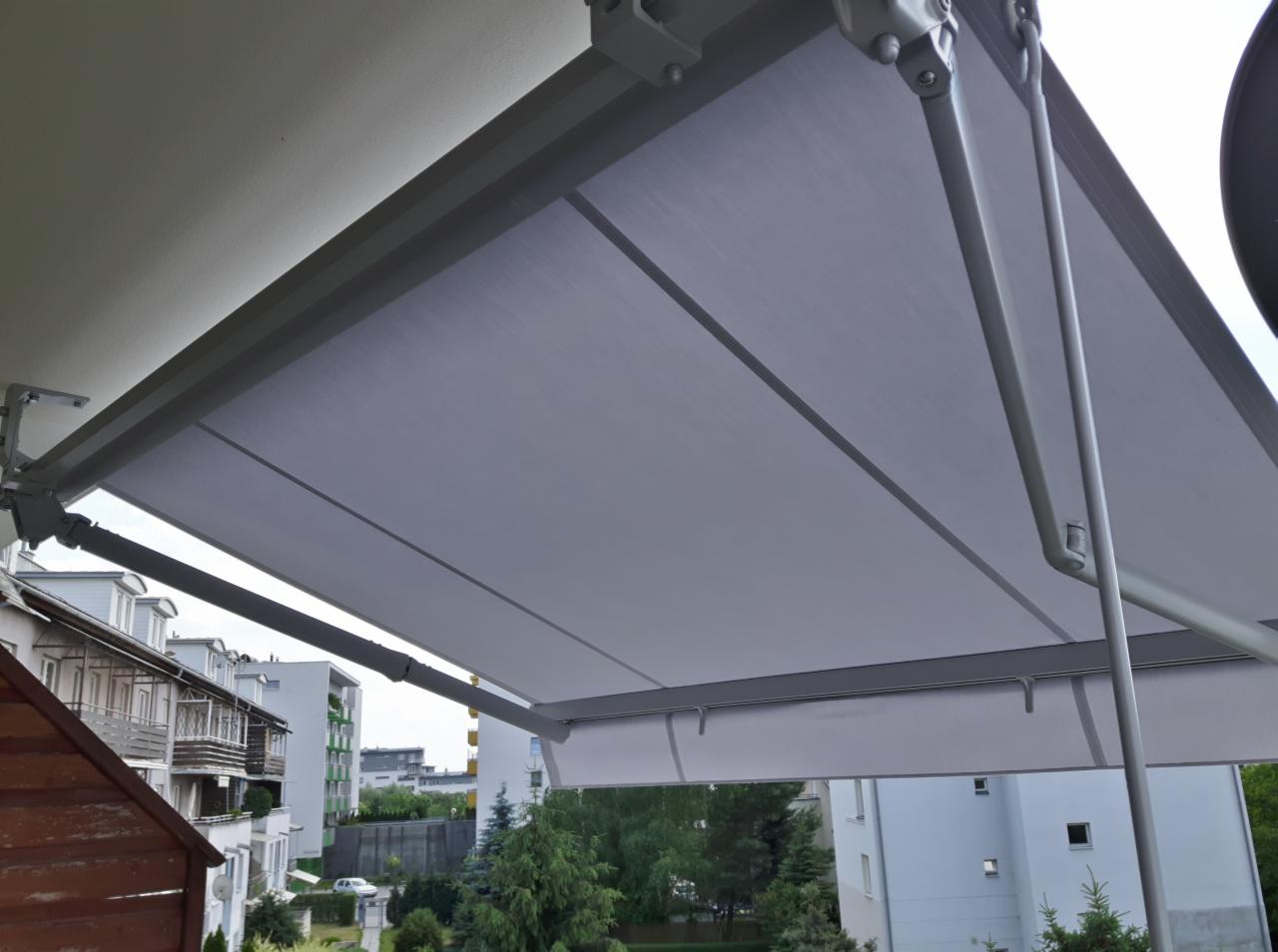 Szara, rozłożona osłona przeciwsłoneczna nad balkonem, widok z dołu na tle budynków mieszkalnych i zieleni.