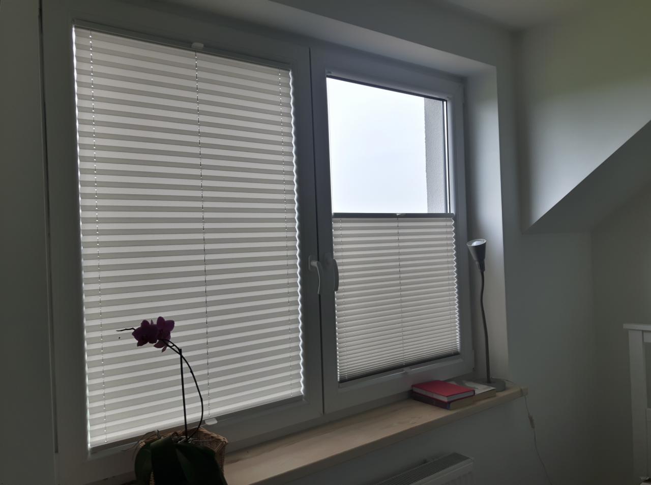 Okno z zamontowanymi plisami w kolorze szarym, obok storczyka i lampki, widok z wnętrza pokoju.